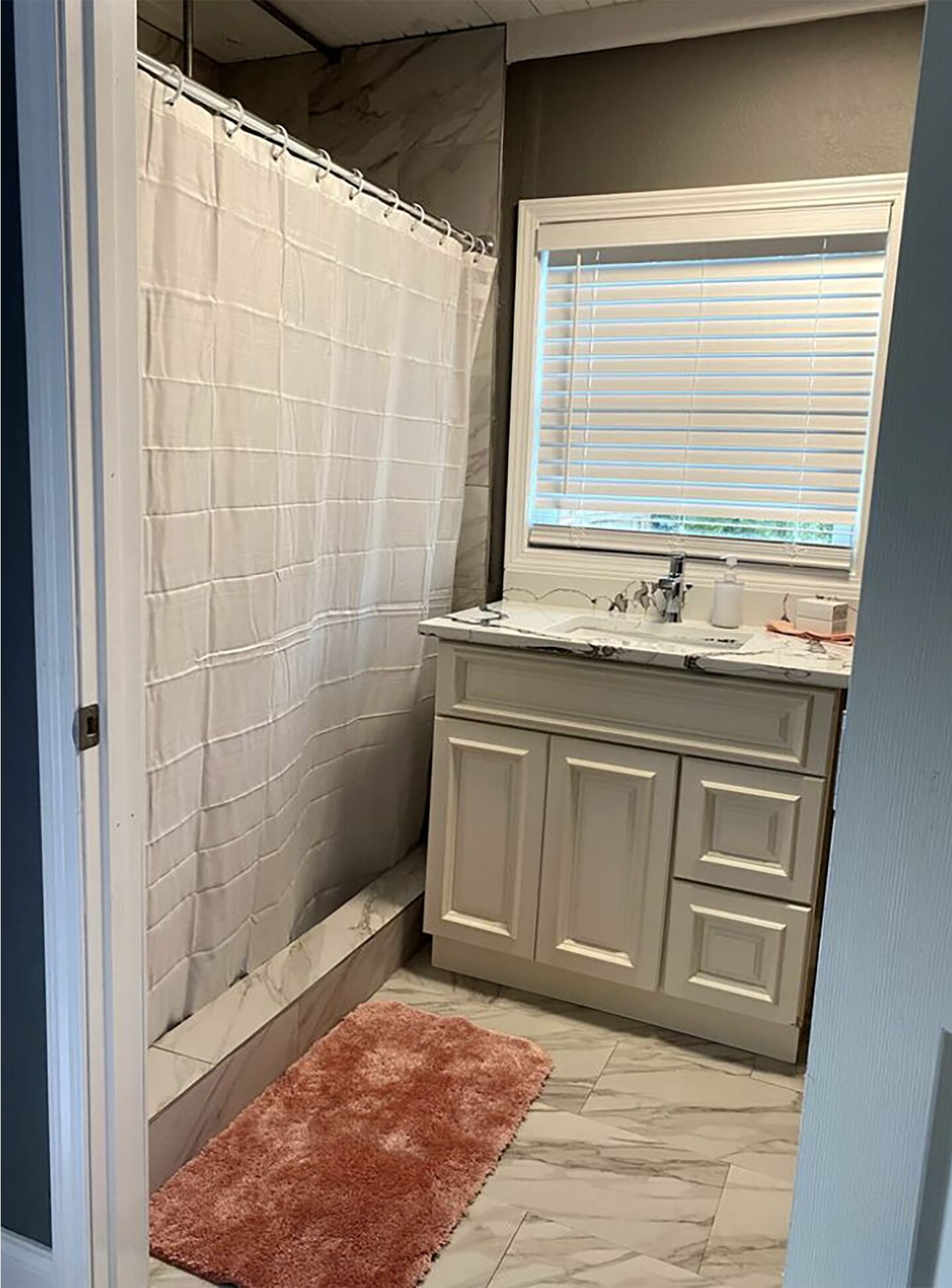 A bathroom with a shower curtain, a window with blinds, a sink, and cabinets.