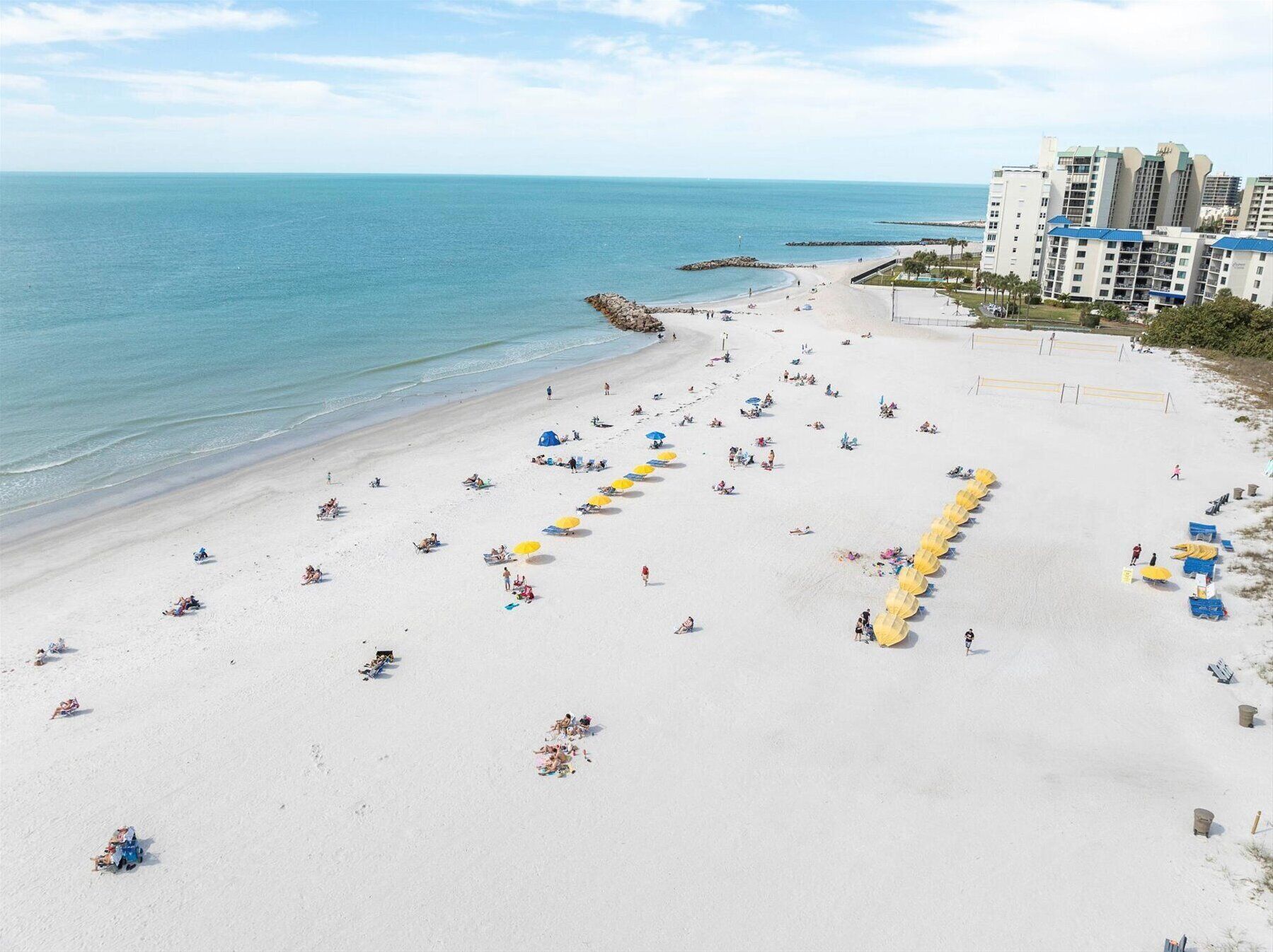A beach with rows of umbrellas and sun loungers, a rocky breakwater, and a hotel in the background.