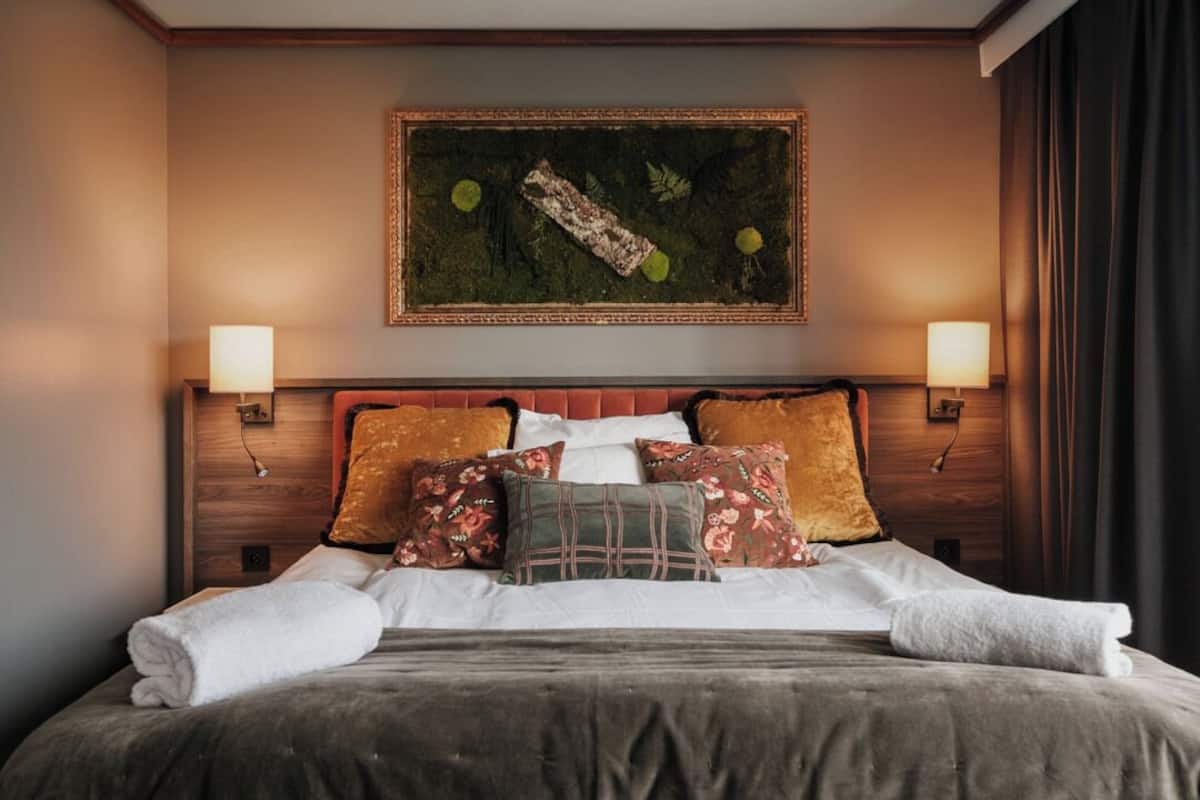 A neatly made bed with a dark bedspread and white linens, flanked by two wall-mounted lamps and a framed artwork above.