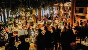A crowded bar area with people standing and sitting, a bartender at the bar, and various bottles and glasses on shelves. - Villa Foresta (Lidingo)