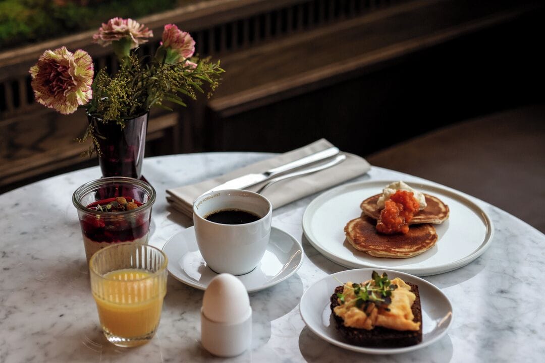 A breakfast setting with a coffee cup, juice, a small egg, pancakes with red sauce, and a dessert on a marble table.
