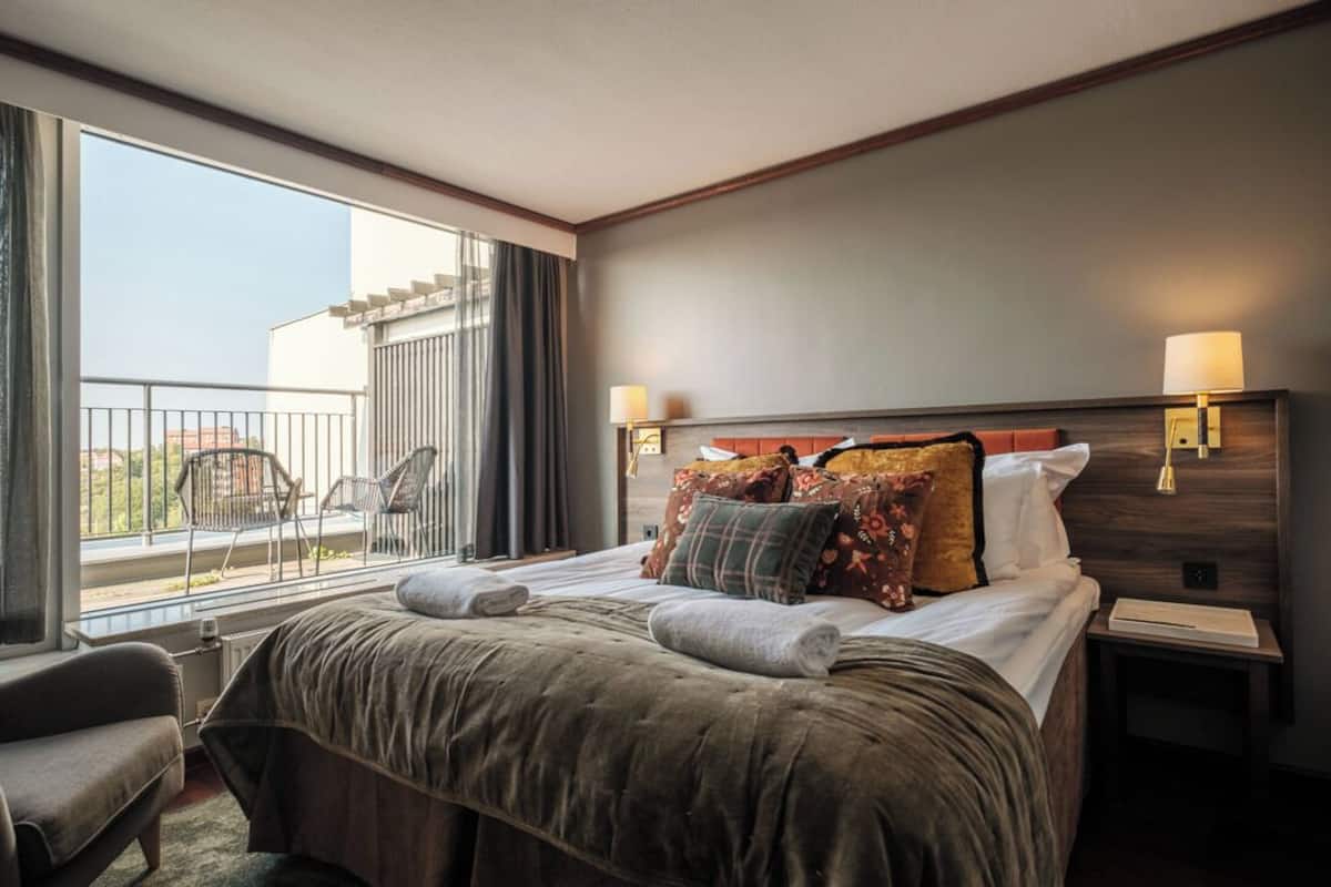 A hotel room with a large bed, two bedside lamps, a nightstand, a chair, and a balcony with chairs.