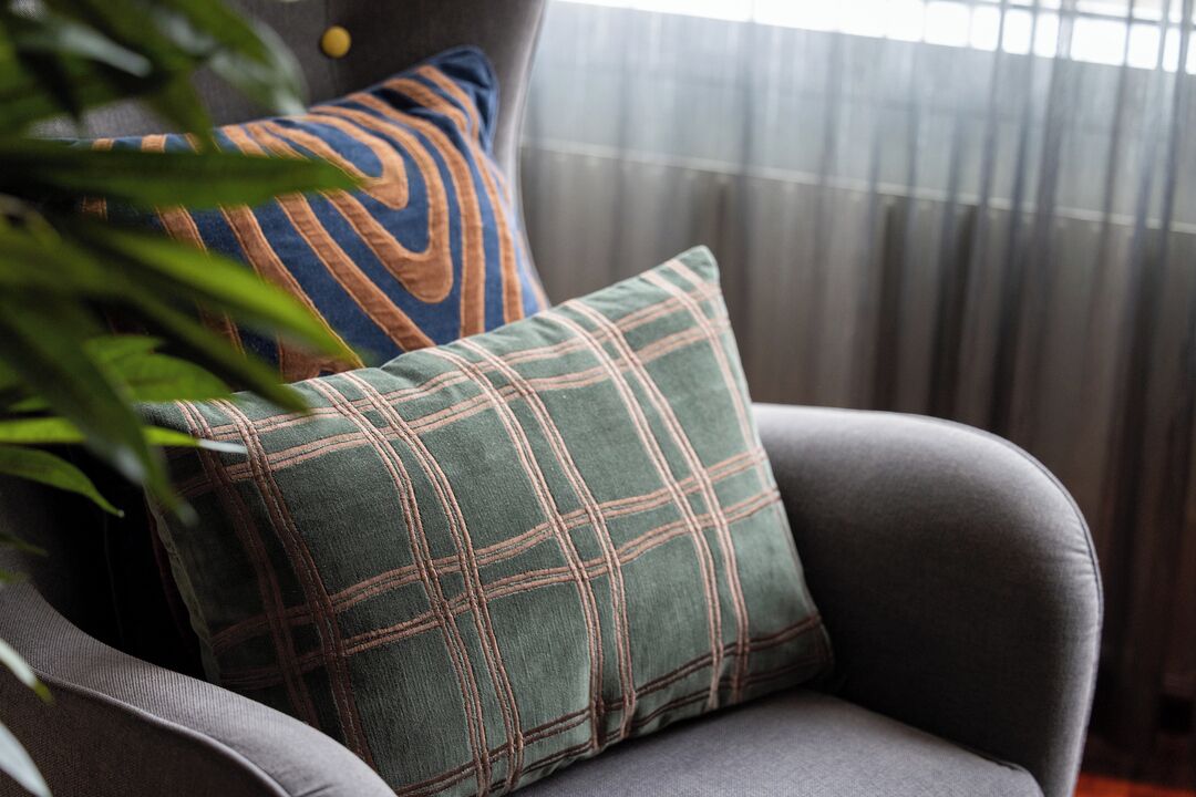 A grey armchair with a plaid pillow and a striped pillow in a room with a window and curtains.