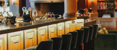 Zona de bar con una barra larga, varios taburetes, lámparas colgantes y una estantería al fondo.
