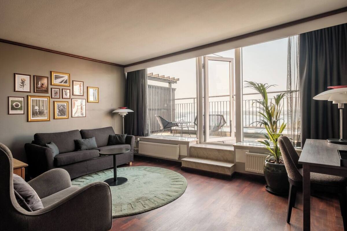 A modern living room with a sofa, armchair, and dining table. There are framed pictures on the wall, a potted plant, and a view of the outside balcony.