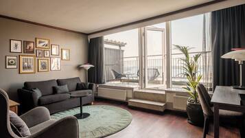 A modern living room with a sofa, armchair, and dining table. There are framed pictures on the wall, a potted plant, and a view of the outside balcony.