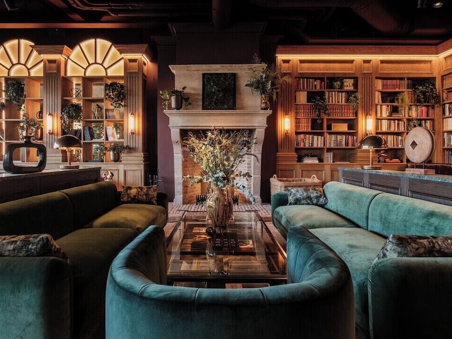 A cozy library with green sofas, a fireplace, and bookshelves filled with books.