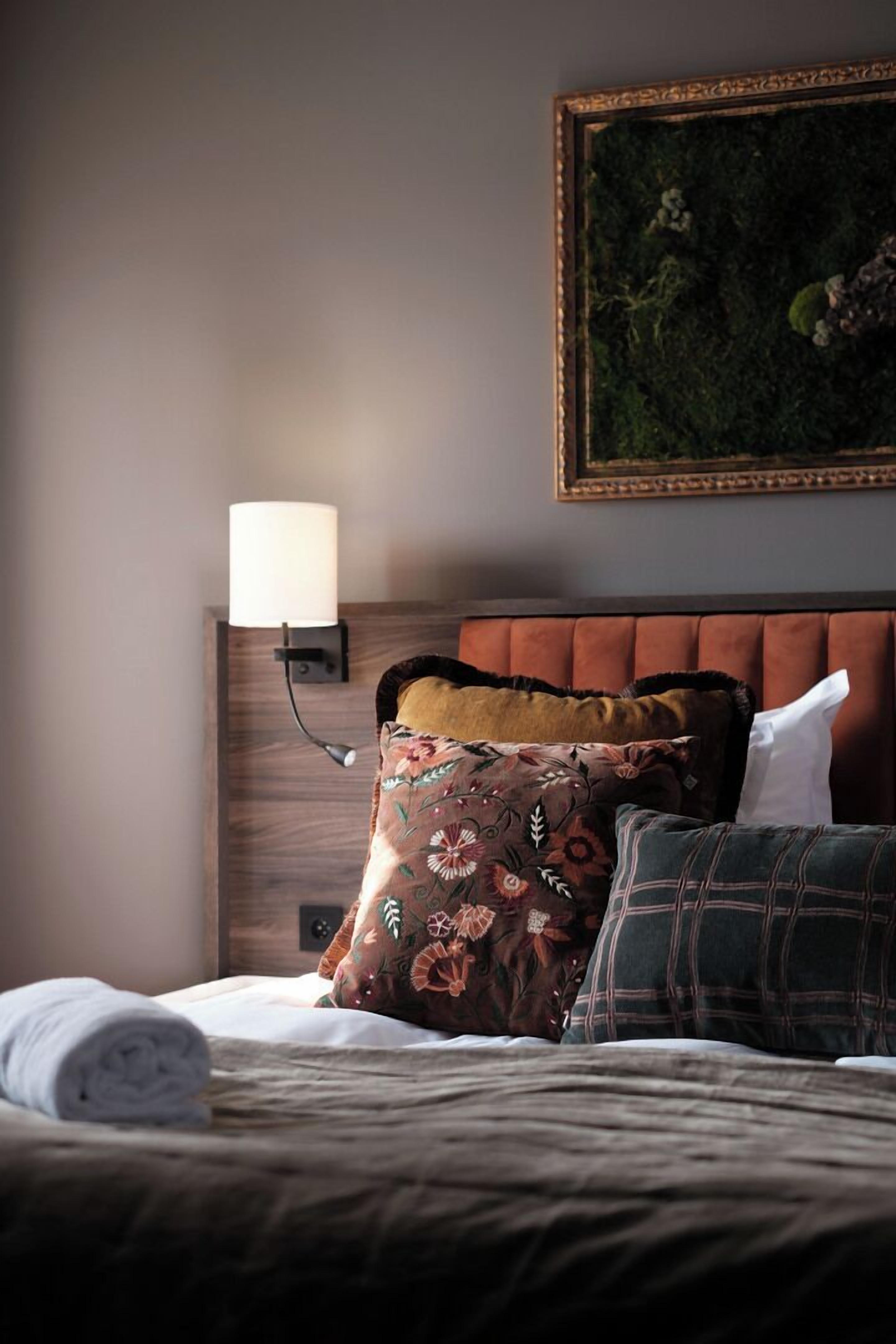 A neatly made bed with a floral-patterned pillow, a plaid pillow, and a white pillow. A wall-mounted lamp is visible on the left side of the bed.