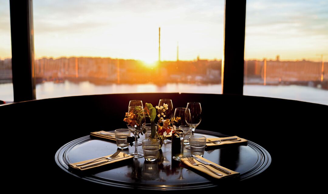 Ein Tisch für vier Personen gedeckt, mit Blumen, Gläsern und Besteck, mit Blick auf ein Gewässer und eine Skyline bei Sonnenuntergang.