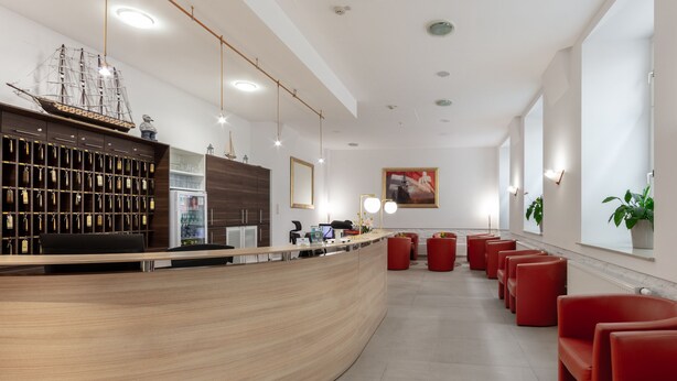 A modern reception area with a curved wooden counter, a ship model, and red seating. - Hotel Admiral (Vienna)