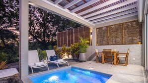 Zona de piscina exterior con sillones de descanso, una mesa y plantas en macetas.
