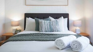 A neatly made bed with a grey knitted blanket, white pillows, and rolled white towels on a white bedspread. There are two bedside lamps on wooden nightstands, and a framed abstract painting hangs above the bed. - Sunset Haven 4-Bd Retreat Milton, Steps to Stadium (Petrie Terrace)