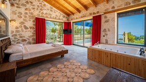 A bedroom with a stone wall, wooden ceiling, a bed, a large window, and a bathroom with a bathtub. - Kaşın en gözdesi taş villalar  (kas)