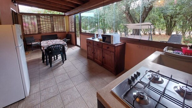 Cozinha com fogão a gás, armários de madeira, mesa de jantar com cadeiras e vista para árvores através de grandes janelas.