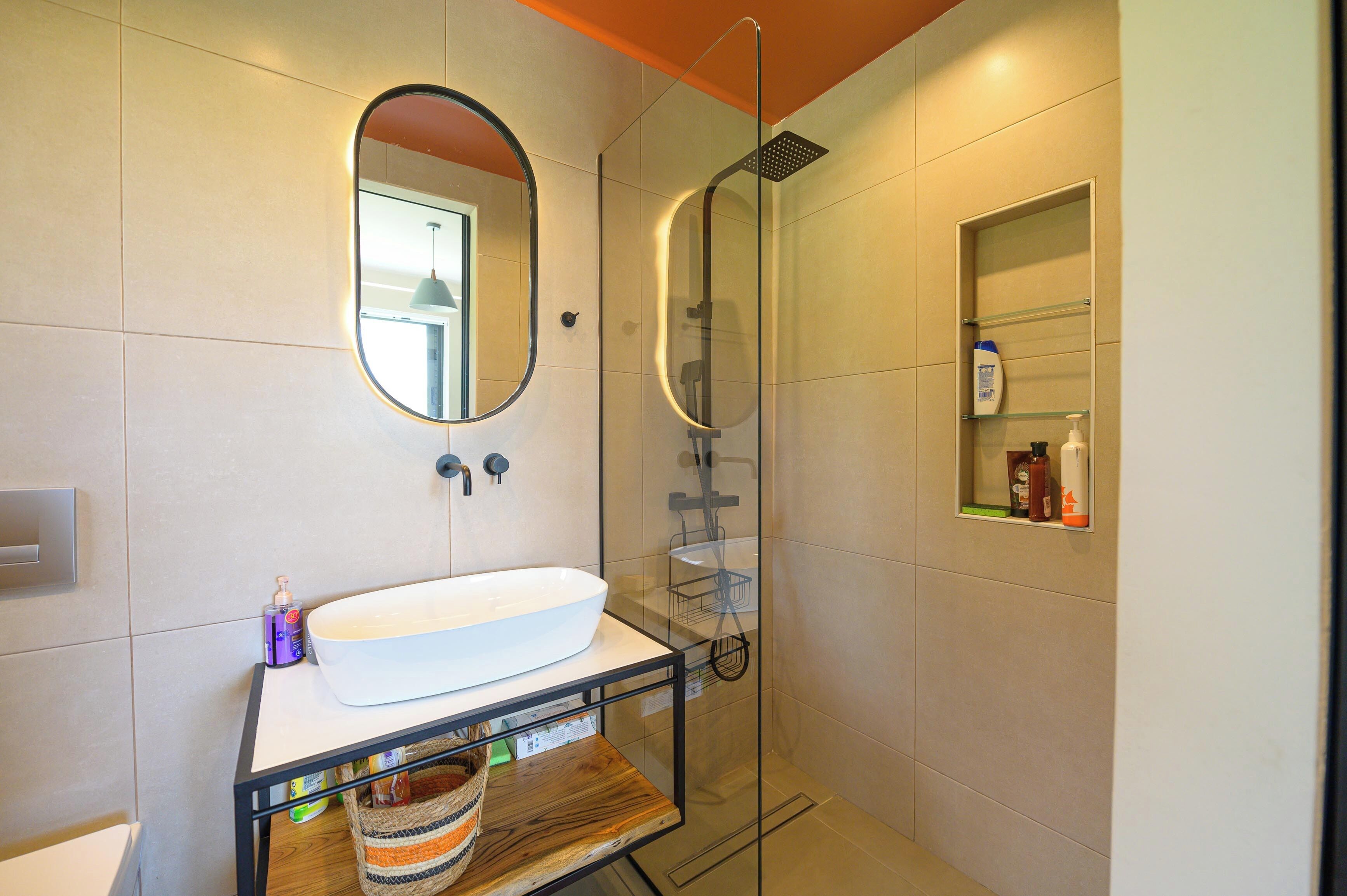 A modern bathroom with a large oval mirror, a white sink, and a glass-enclosed shower.