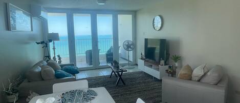 A living room with a sofa, a TV, a dining table with a floral centerpiece, and a balcony with a view.
