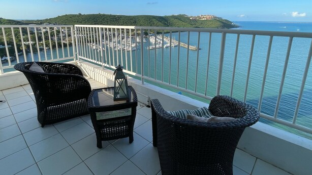 A balcony with wicker furniture, a lantern, and a view of a marina and hills. - Ocean View 2 Bed 1 Bath 2 Parkings Pool (Fajardo)