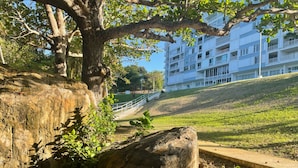 A large rock formation with a tree in the foreground, a grassy slope, and a white building with balconies in the background. - Ocean View 2 Bed 1 Bath 2 Parkings Pool (Fajardo)