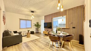 A modern living room with a sofa, a dining table with chairs, a TV mounted on the wall, and a large window.