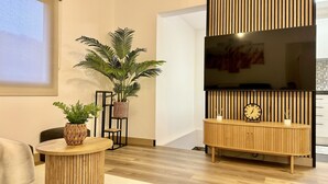 Una sala de estar moderna con piso de madera, una mesa redonda de madera con una planta, un soporte de metal negro, una cesta tejida, una consola de madera con un reloj y un televisor de pantalla plana grande montado en una pared de paneles de madera.