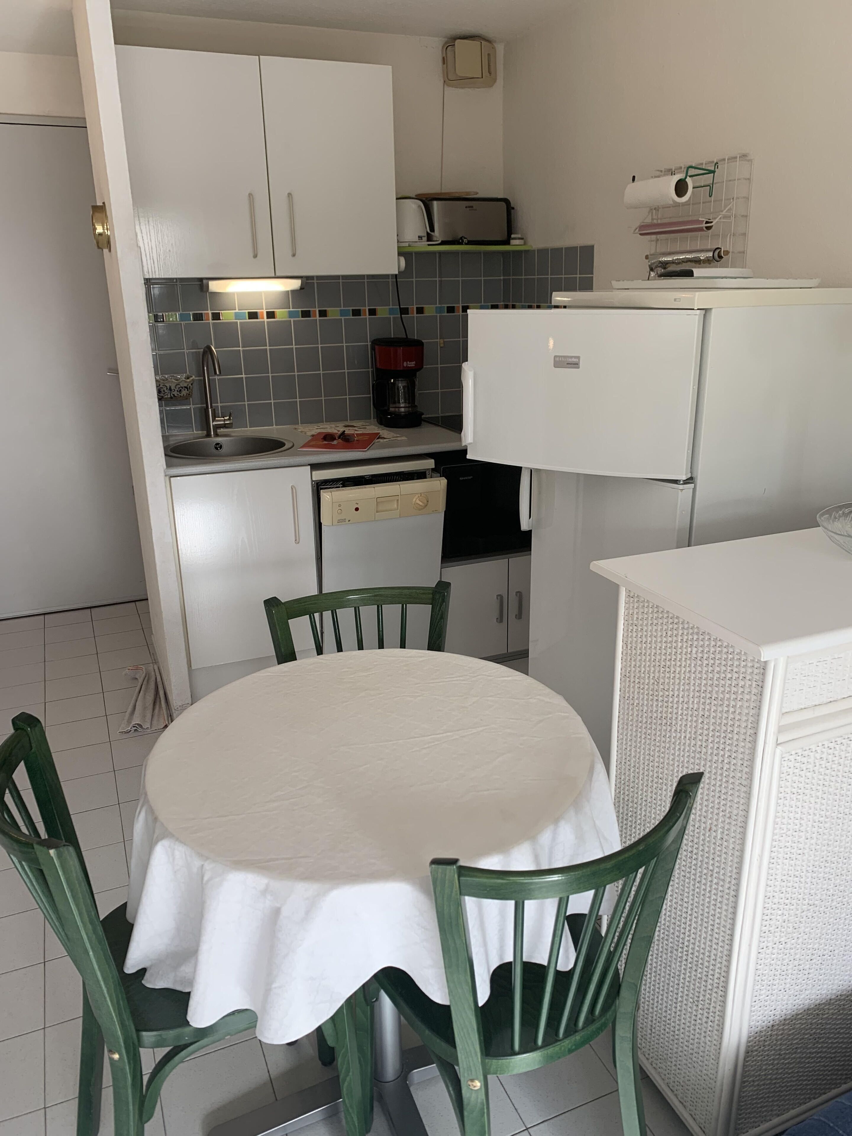 Eine kleine Küche mit einem runden Tisch und grünen Stühlen, einer Kaffeemaschine und einem Kühlschrank.