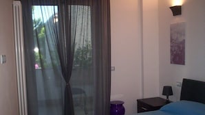 Camera d'albergo con un letto, un comodino, una lampada, una finestra con tende, un termosifone e un punto luce a parete.