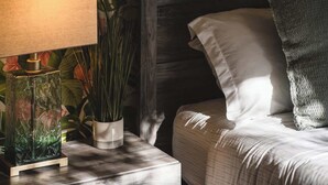 A neatly made bed with a white pillow and a dark gray throw pillow, a bedside table with a lamp and a decorative vase, and a plant in a white pot. - Paradise Haven-95 walk score-Dedicated Workspaces (Portland)