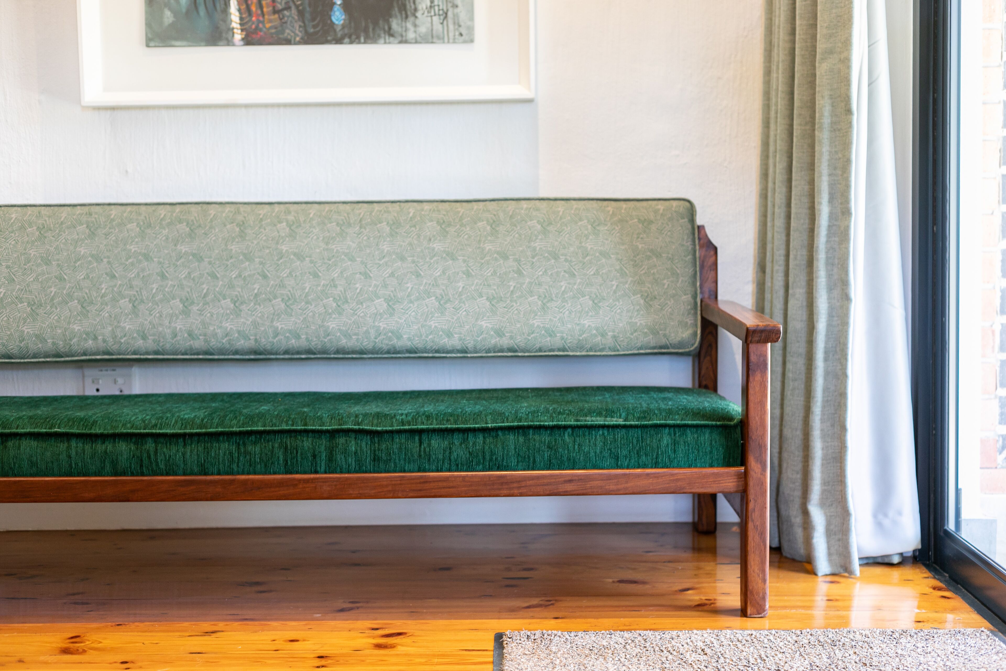 A wooden bench with a green cushion against a white wall, a framed artwork above it, and a window with curtains to the right.