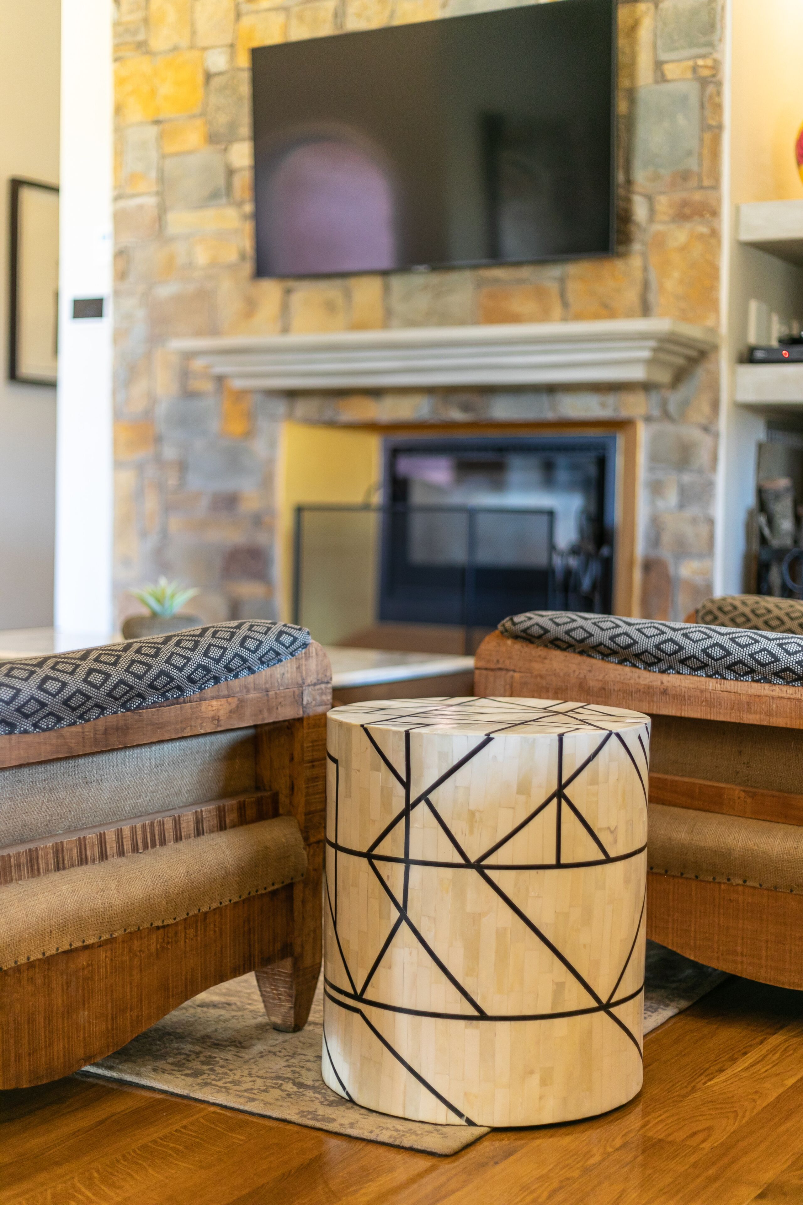 A cozy living room with a stone fireplace, a flat-screen TV mounted above it, and two patterned ottomans in front of a wooden sofa.