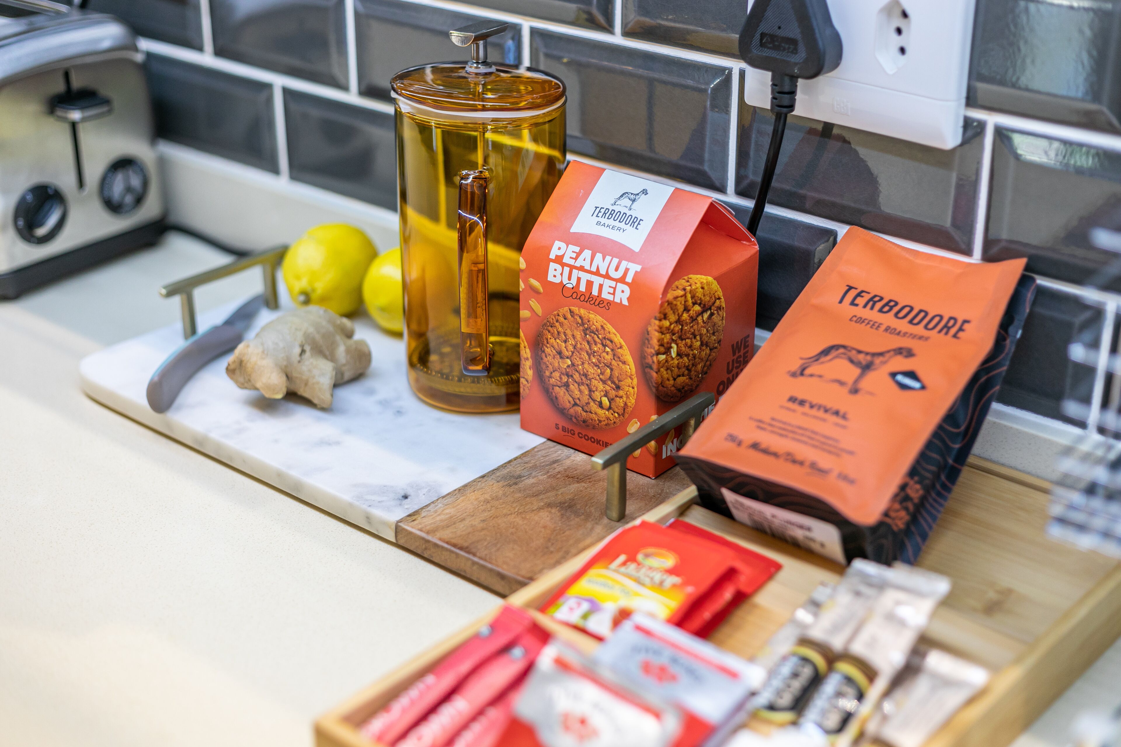 Una encimera de cocina con una cafetera, una caja de galletas de mantequilla de cacahuete, un paquete de café Terbodeore, limones, jengibre y varios utensilios de cocina.