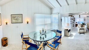 Un'area pranzo con tavolo in vetro e sedie blu, interni bianchi con travi in legno e vista su un soggiorno con divano e camino.