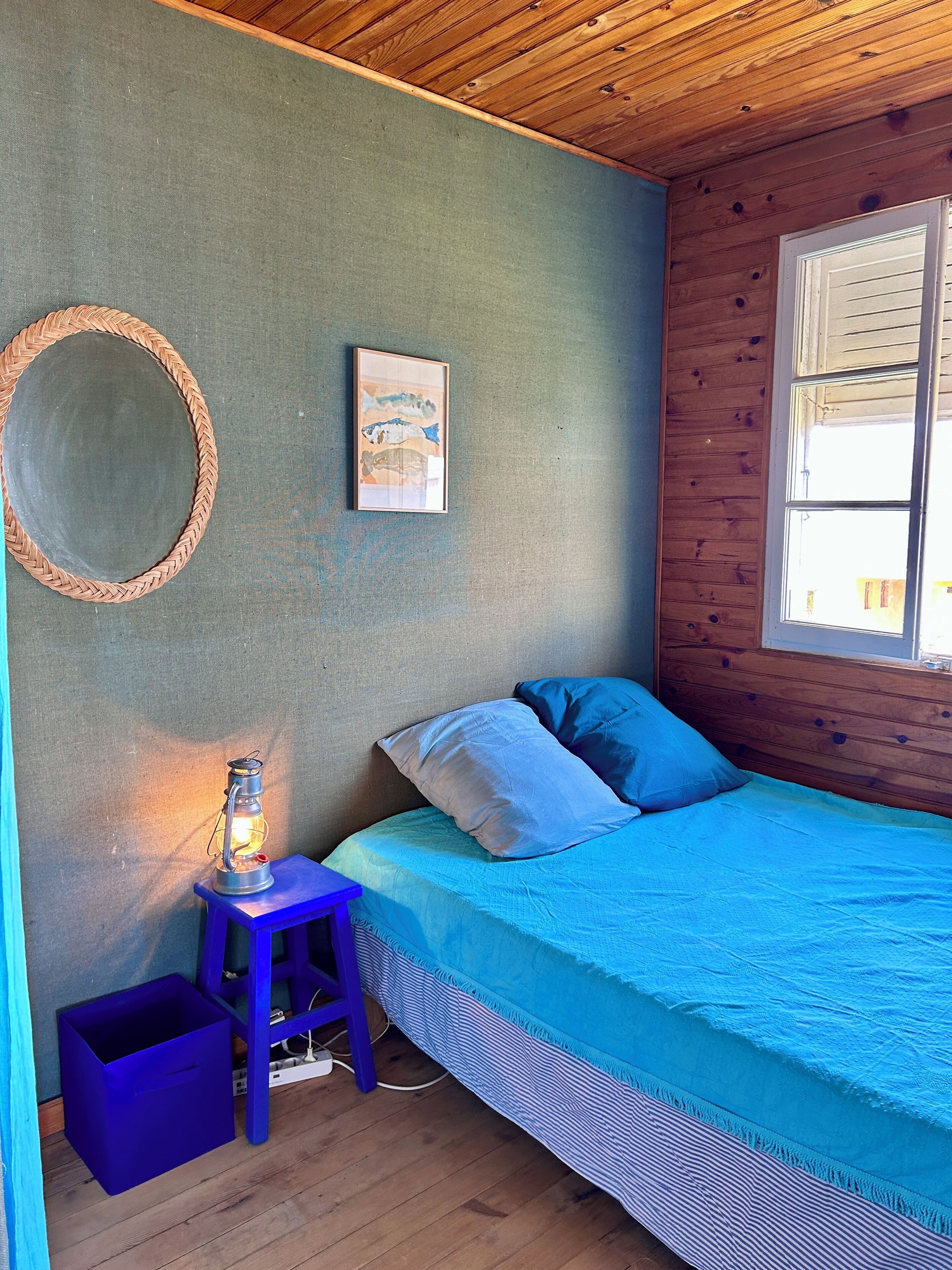 A bedroom with a bed, a small stool, a lantern, a framed picture, and a window.
