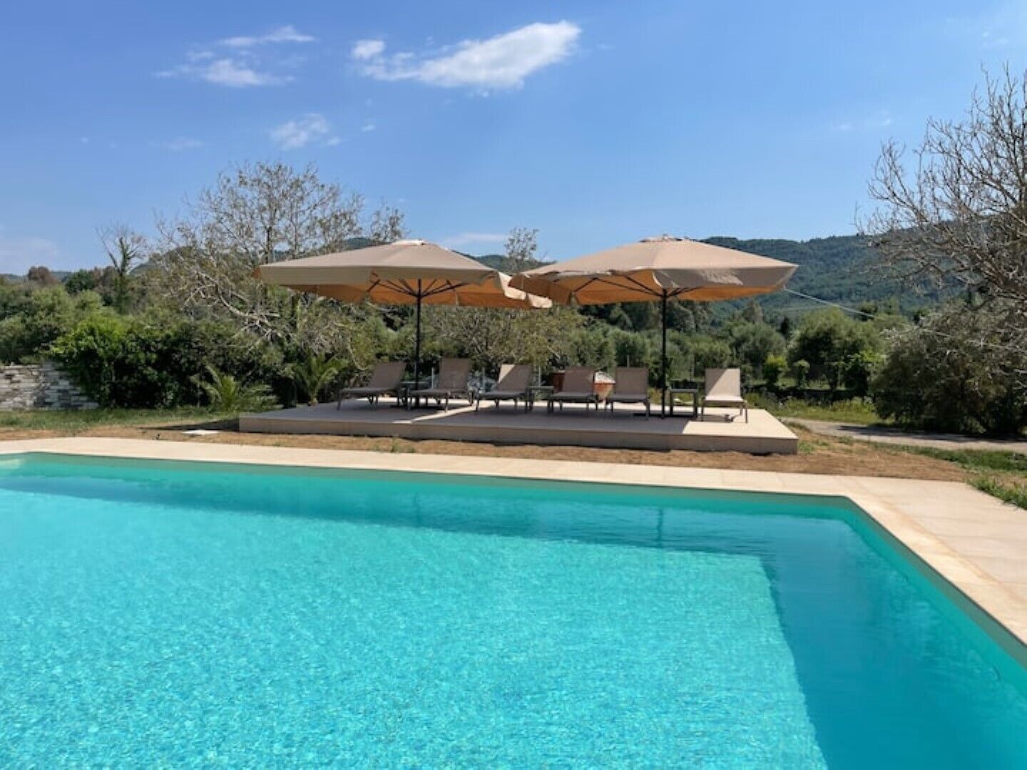 Une piscine à l’eau d’un bleu cristallin, entourée de chaises longues et de parasols, avec en arrière-plan un paysage de verdure composé d’arbres et de collines.
