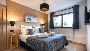 Een slaapkamer met een houten wand, een bed met een geruit kleed, twee bedlampen, een ingelijste foto, een raam met gordijnen en een radiator.