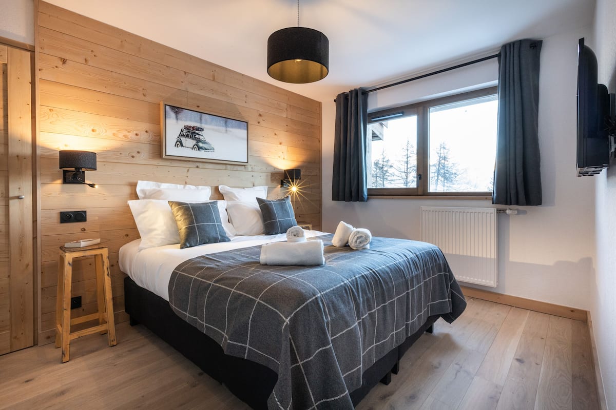A bedroom with a wooden wall, a bed with a checkered blanket, two bedside lamps, a framed picture, a window with curtains, and a radiator.