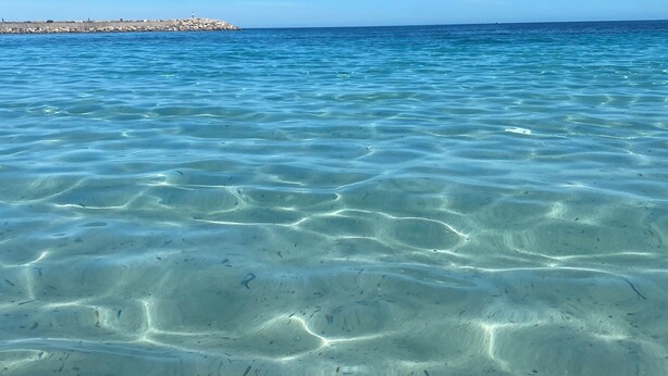 Clear shallow waters with visible sand patterns and a distant rocky shoreline.