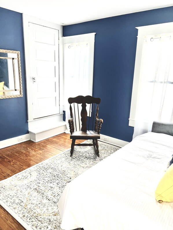 A bedroom with a wooden floor, a large bed, a chair, and two windows with white curtains. - Vintage Retreat: 5 minute walk to downtown, Naval Academy, Stadium and more.
(Annapolis)