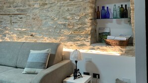 A living room with a stone wall, a grey sofa, a small white table with a lamp, and a shelf with bottles. - Censo Guest House : are you ready for your next adventure? (Marsaglia)