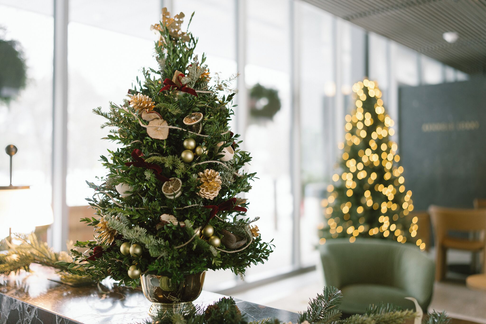Un albero di Natale decorato con ornamenti e verde, con un albero di Natale illuminato sullo sfondo e una poltrona verde.