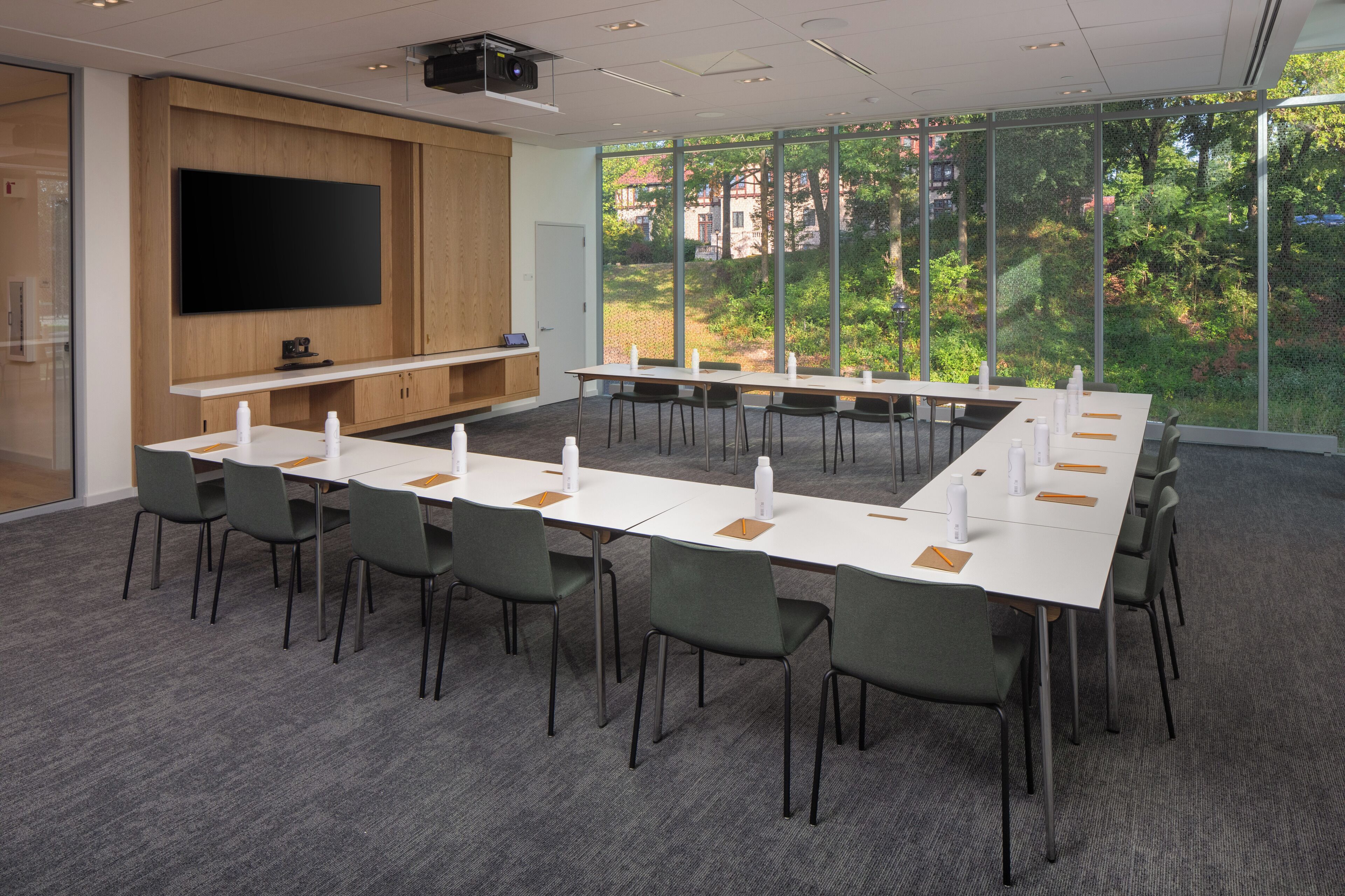 A conference room with a long table, chairs, a flat-screen TV, and large windows with a view of greenery.