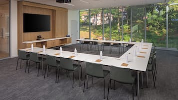 A conference room with a long table, chairs, a flat-screen TV, and large windows with a view of greenery.