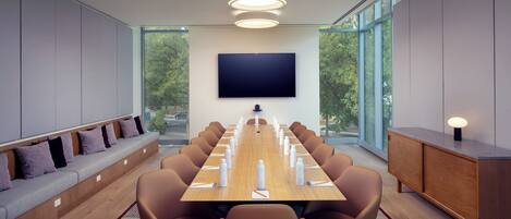 A conference room with a long table, brown chairs, a flat-screen TV, and a seating area with cushions.