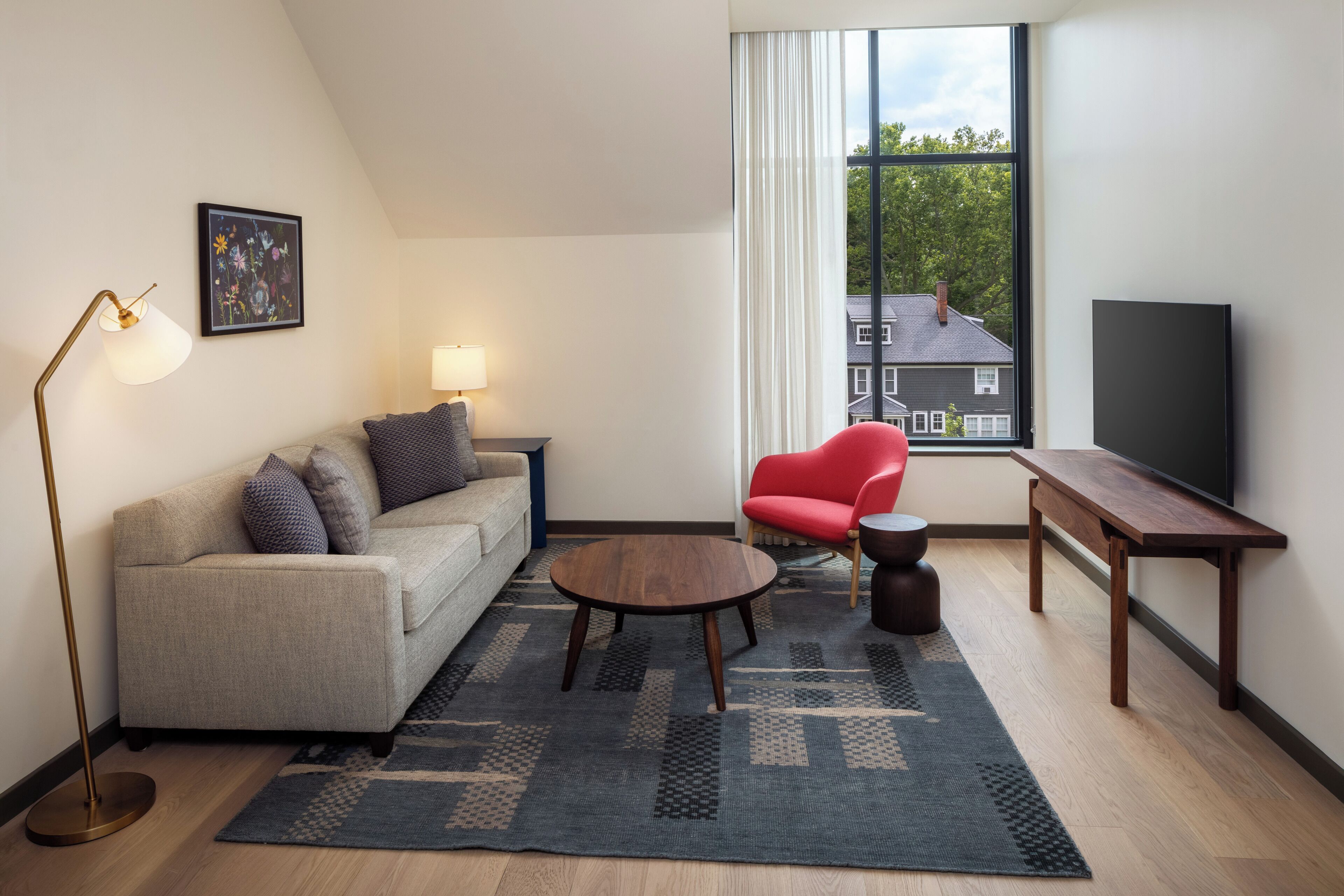 Ein modernes Wohnzimmer mit einer Couch, einem roten Sessel, einem Couchtisch, einer Stehlampe, einem Fernseher und einem großen Fenster mit Blick auf ein Haus.