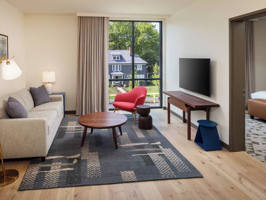 A modern living room with a sofa, a red chair, a round coffee table, a TV, and a large window with a view of a house.