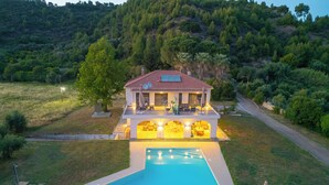 A two-story house with a swimming pool, surrounded by a landscaped garden and a hilly backdrop.