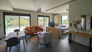 A modern living space with a dining area, sofa, TV, kitchen, and large windows offering a view of the outdoors.