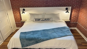 A neatly made bed with a blue blanket, white pillows, and a 'STAY awhile' pillow. - The Apalachicola Train Depot (Apalachicola)