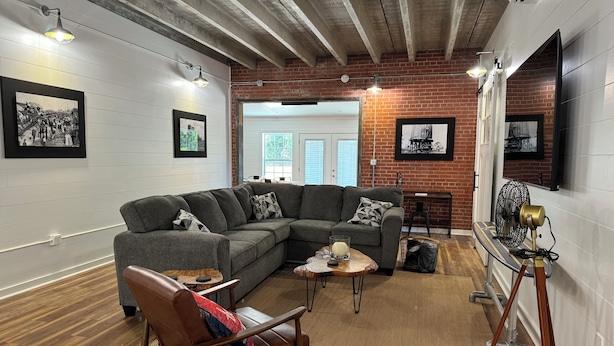A living room with a sectional sofa, a coffee table, and a leather armchair. There is a brick wall, a white door, and a television mounted on the wall. - The Apalachicola Train Depot (Apalachicola)