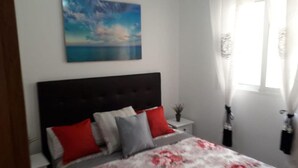 A bedroom with a dark headboard, a bed with red and grey pillows, a floral bedspread, a painting of a seascape, and a window with sheer curtains. - El Campello Deborah. à 10 Minutes à Pied de la Plage et 5 Minutes du Centre (El Campello)
