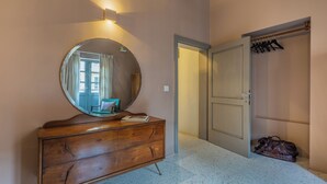 A room with a wooden dresser, a round mirror, and a door leading to a closet with hanging clothes. - Mid-Century Retro Suite (Floriana)